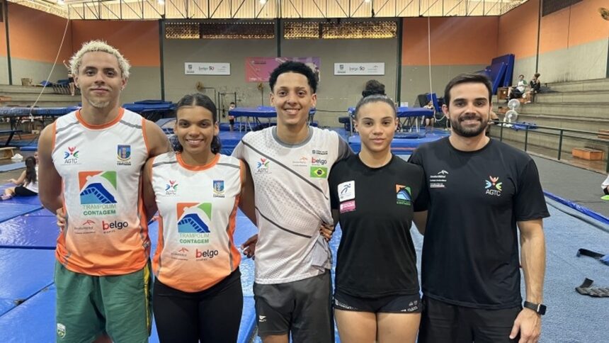 Quatro atletas e um técnico de trampolim sorriem em ginásio com trampolins