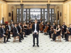 Orquestra com maestro e mais de vinte músicos de cordas em salão clássico com pilares.