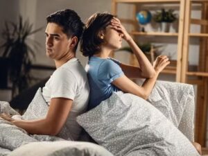 Casal sentado de costas na cama, homem sério, mulher com a mão na testa