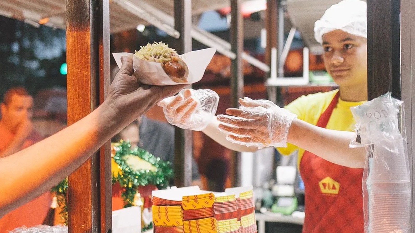 Mão de cliente entregando cachorro-quente com batata palha a vendedor(a) com luvas