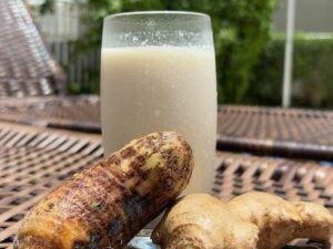 Copo com suco claro, inhame e gengibre sobre textura marrom de vime, ao fundo plantas