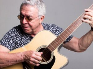 Luiz Carlos Sá, homem idoso de óculos escuros, tocando violão acústico em fundo cinza.