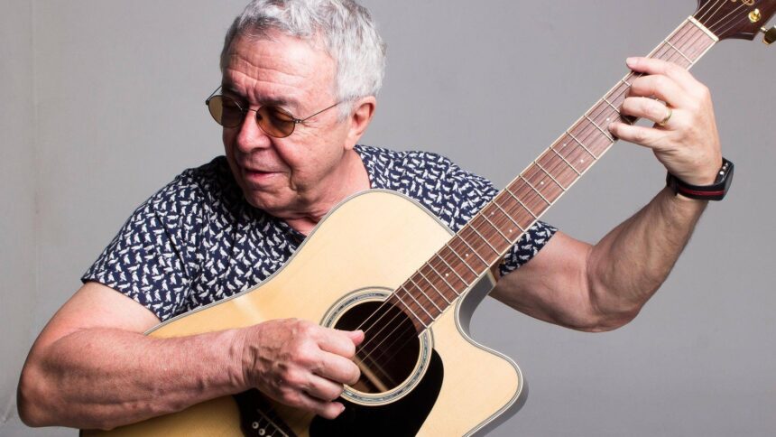 Luiz Carlos Sá, homem idoso de óculos escuros, tocando violão acústico em fundo cinza.