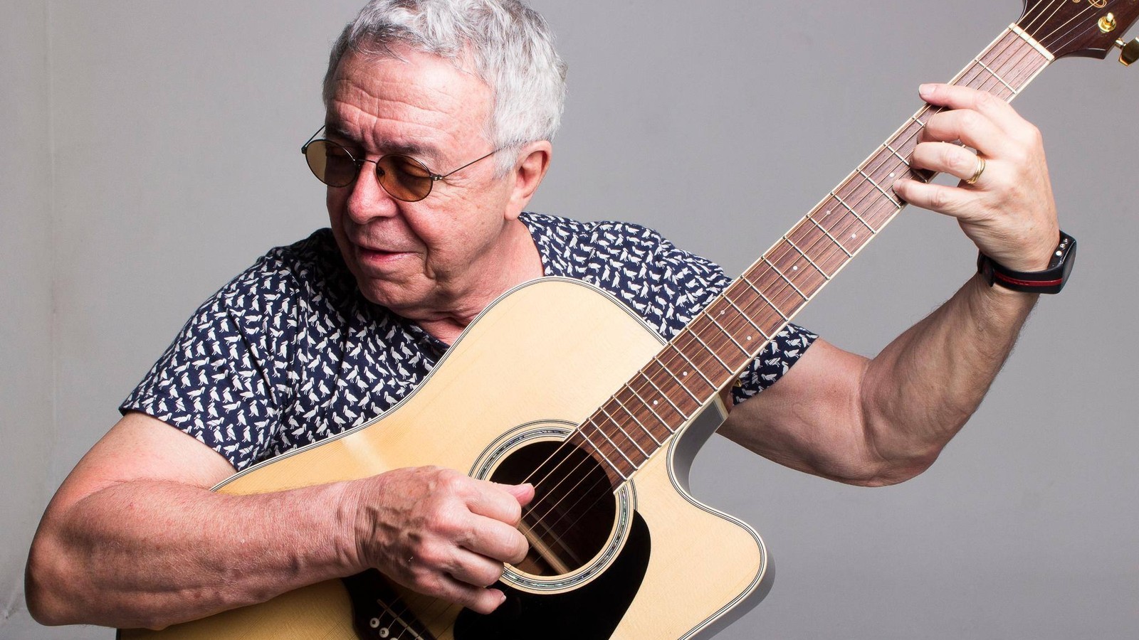 Luiz Carlos Sá, homem idoso de óculos escuros, tocando violão acústico em fundo cinza.