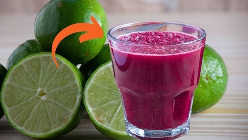 Um copo de suco vermelho escuro ao lado de limões verdes frescos, com uma seta laranja