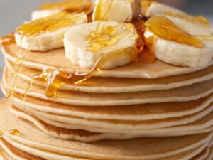 Pilha alta de panquecas com rodelas de banana e calda dourada escorrendo