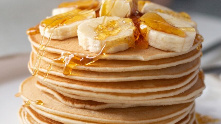 Pilha alta de panquecas com rodelas de banana e calda dourada escorrendo