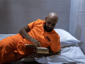 Detento em uniforme laranja D.O.C. com tatuagens lê livro deitado em cela prisional